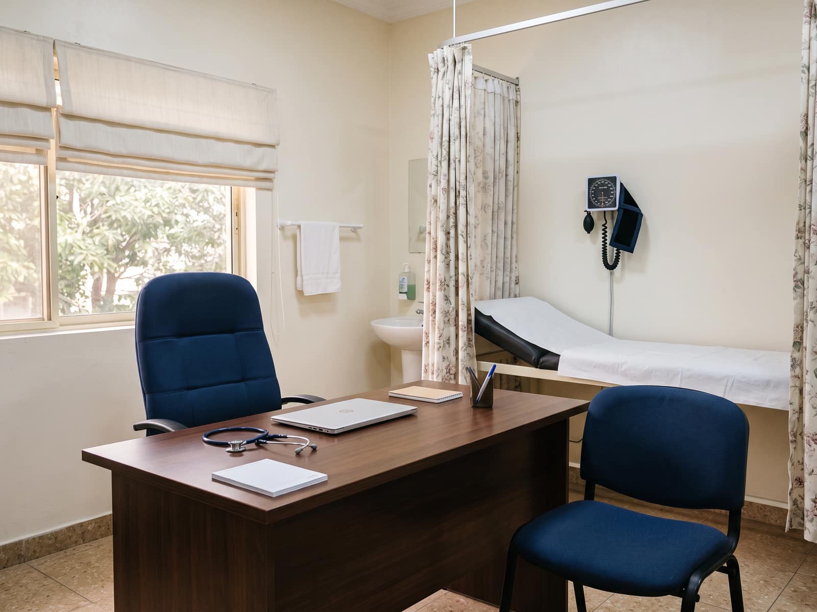 A consulting room with a desk, two chairs, an examination couch and a blood pressure monitor
